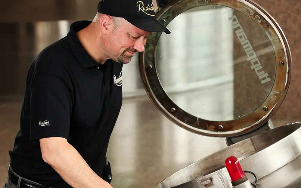 A man in work clothes wearing a Radeberger cap is inspecting brewing equipment at a production facility.