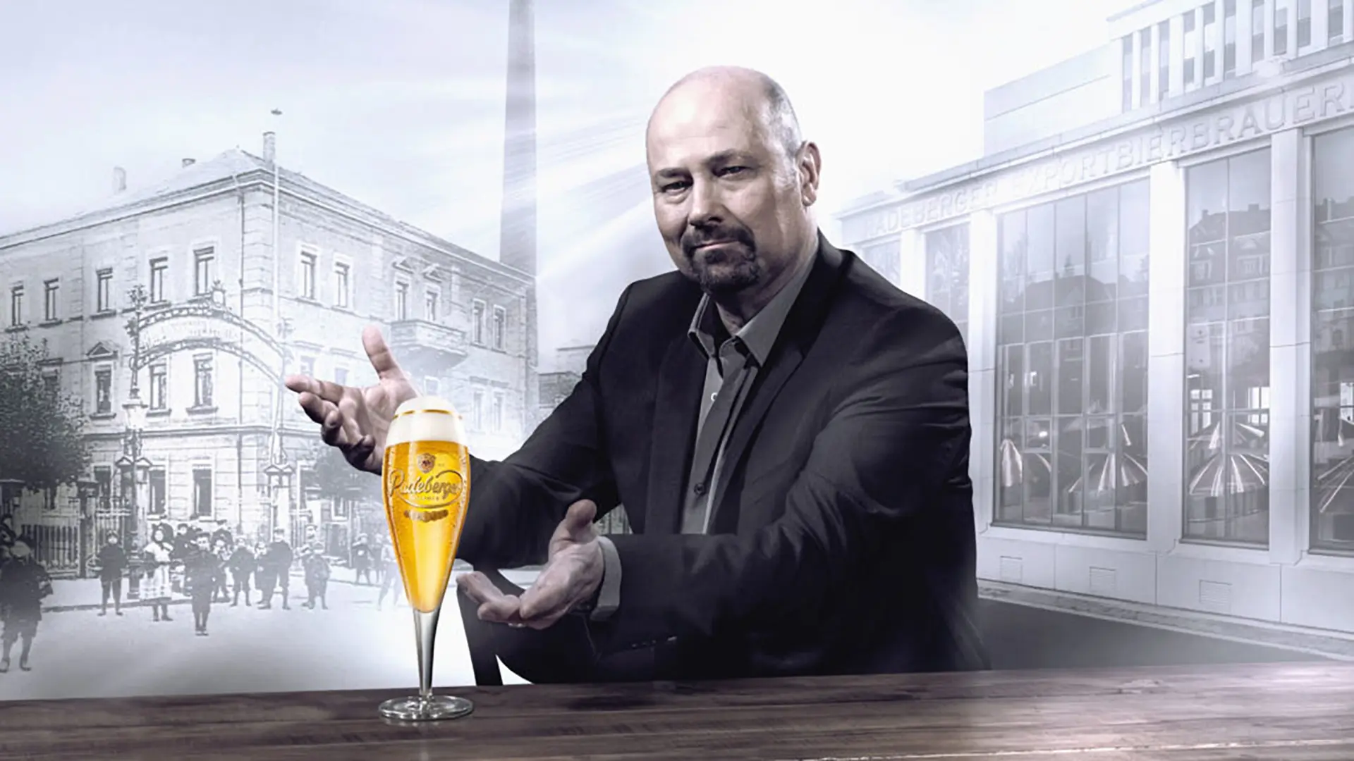 Man in a suit presenting a glass of radeberger beer on a table. Historic and modern brewery buildings in the background.