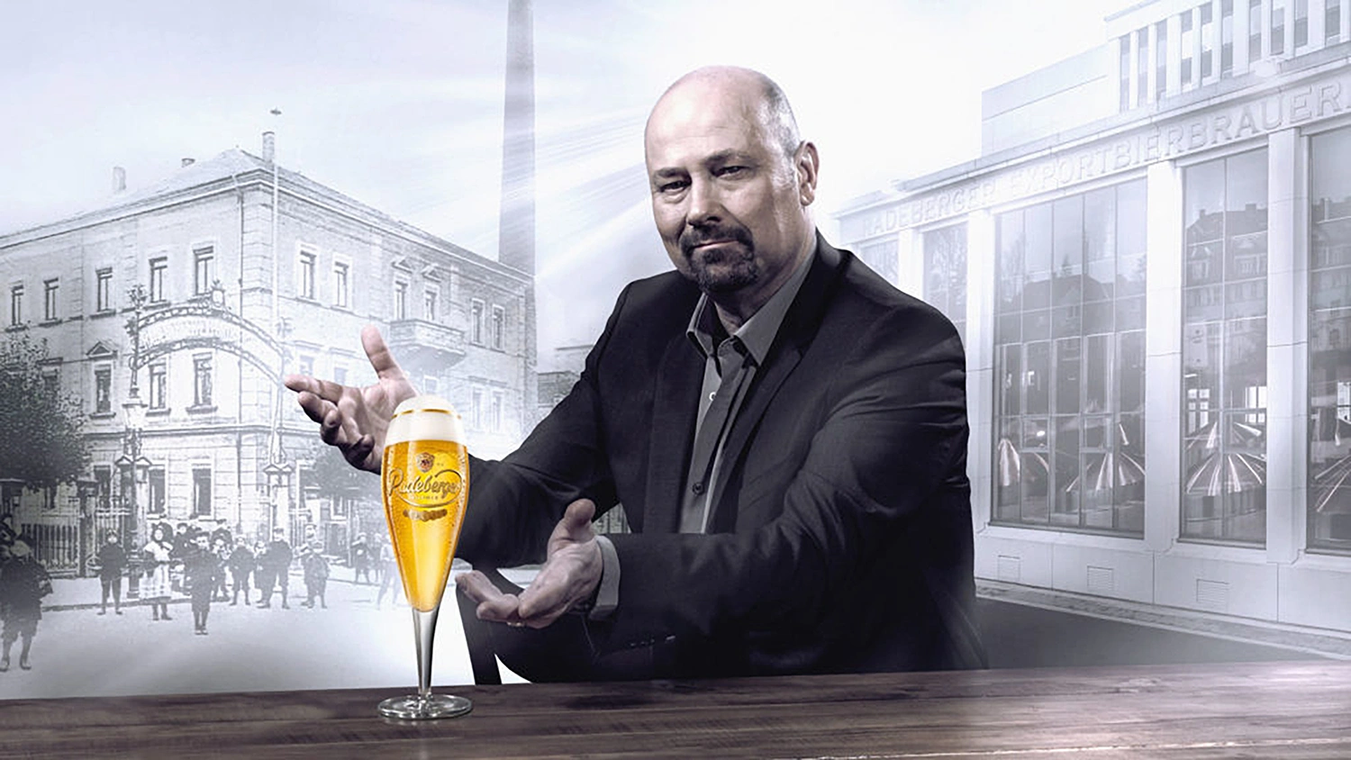 Man in a suit presenting a glass of radeberger beer on a table. Historic and modern brewery buildings in the background.