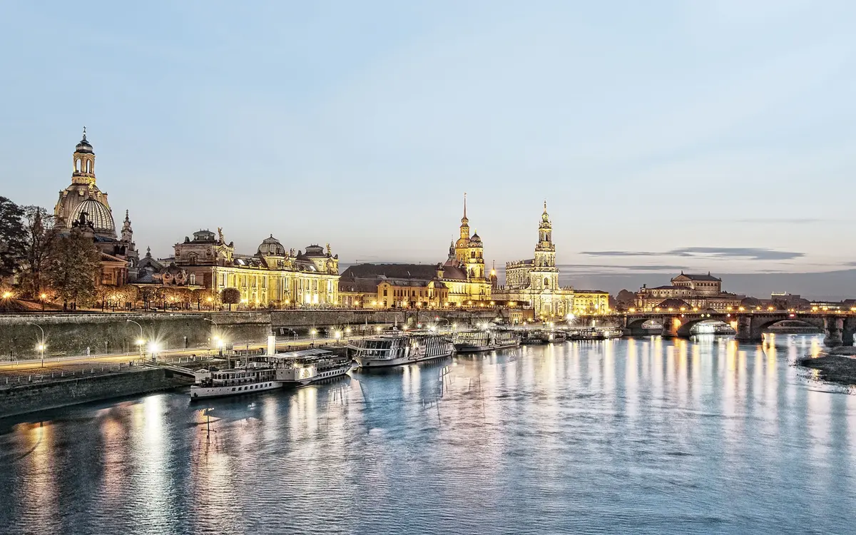 Historic city skyline with illuminated buildings and boats along a river. Soft evening sky forms the background.