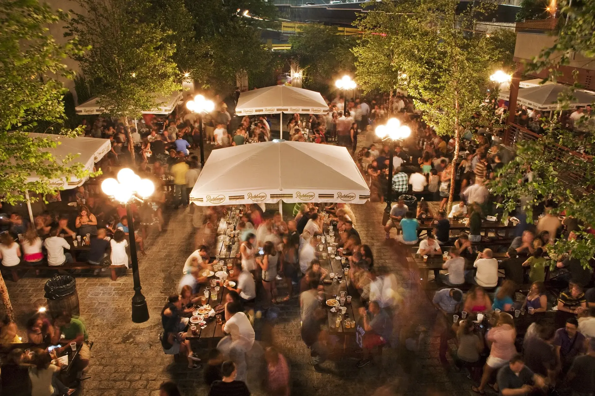 People sit and stand at tables in a beer garden at night. Trees and lamps surround the area.
