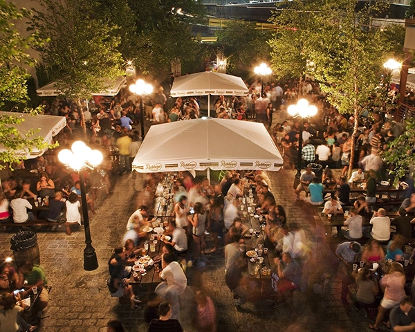 People sit and stand at tables in a beer garden at night. Trees and lamps surround the area.