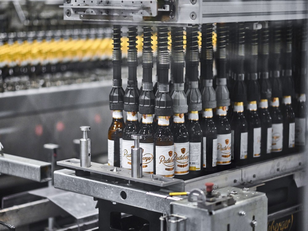 A conveyor belt in a factory is filling and labeling clear bottles with amber liquid.