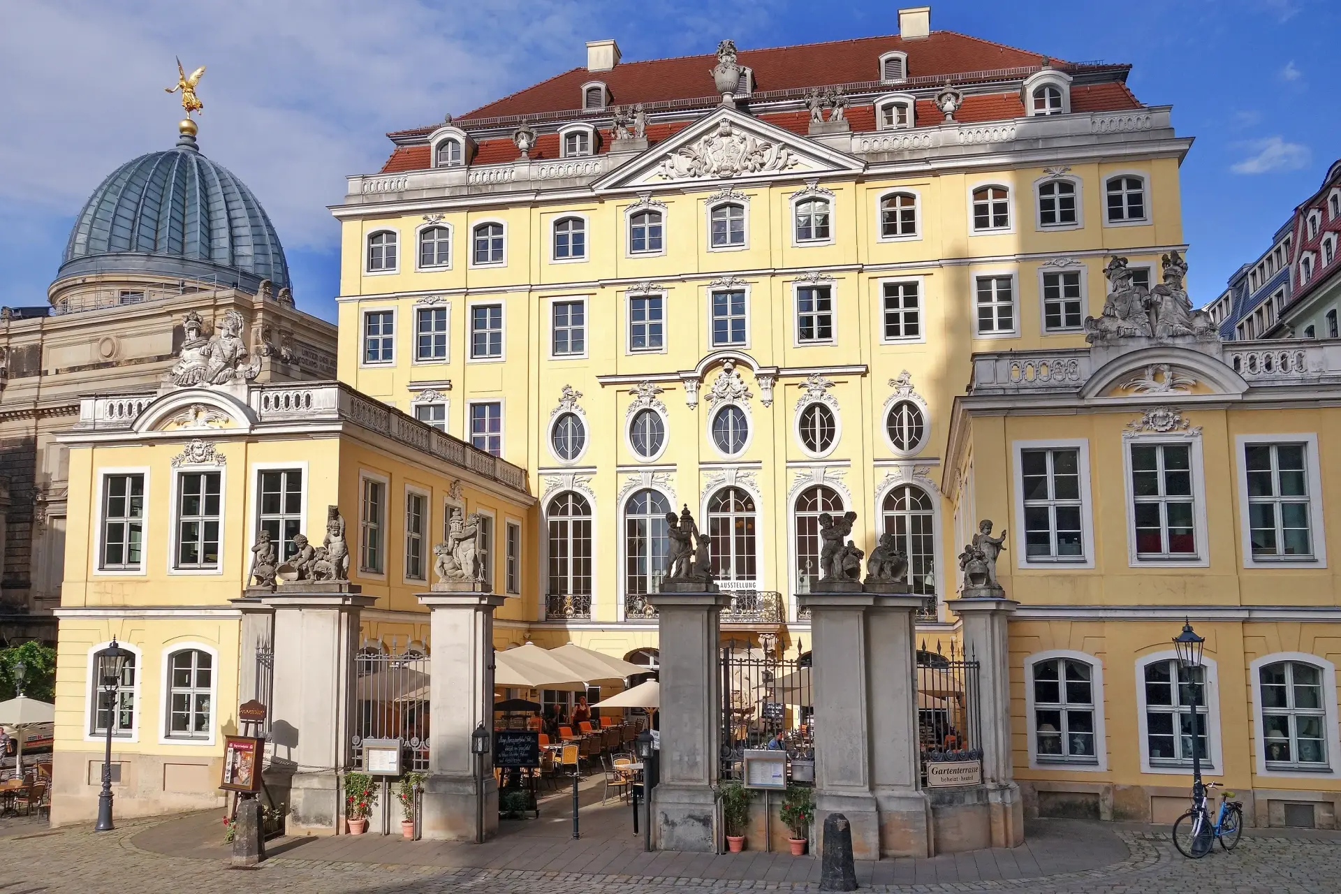 Historic building with statues gate and courtyard seating. Yellow facade with ornate details and blue sky.
