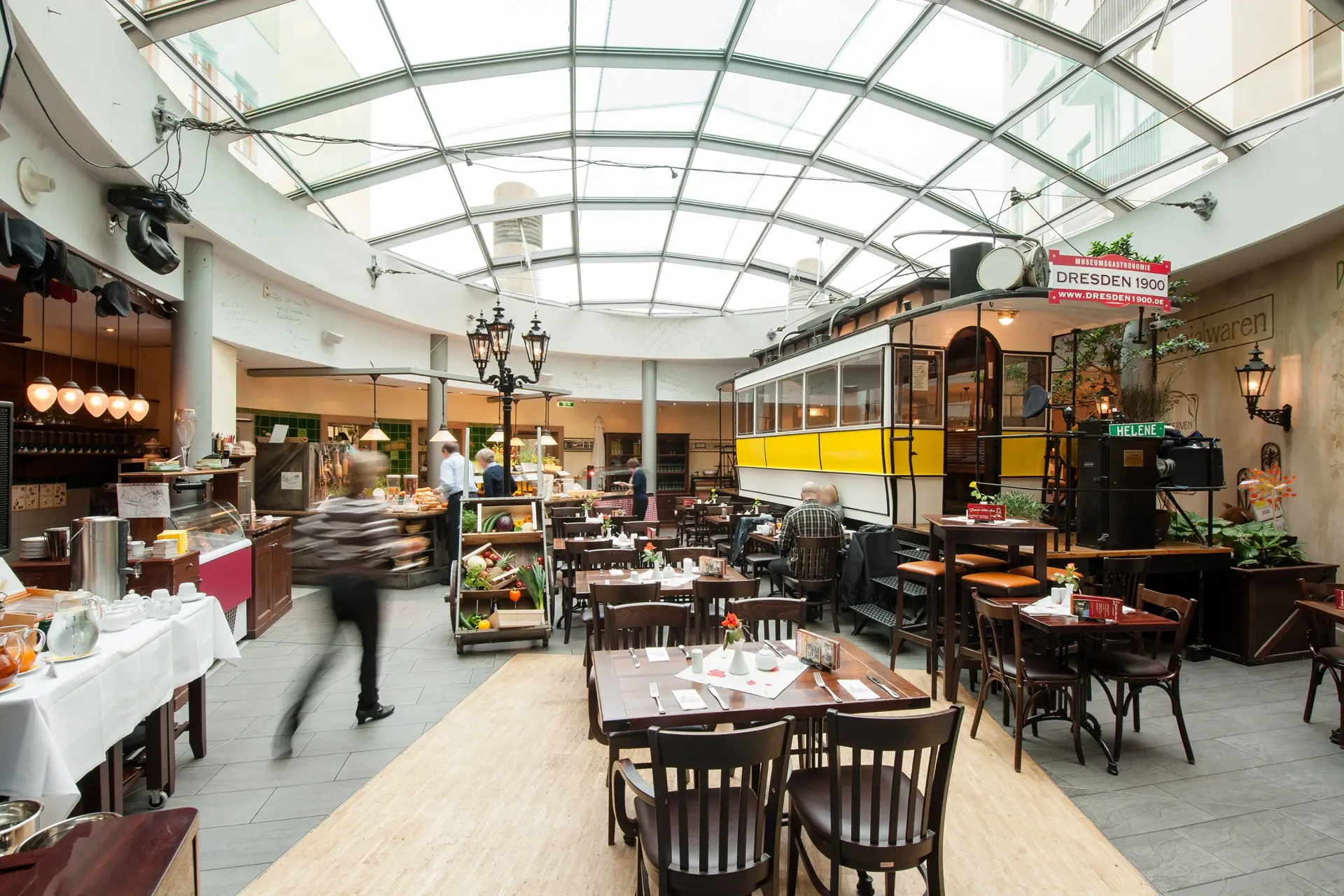 Restaurant interior with tables chairs and historic tram. Glass roof above buffet counters and guests moving through space.