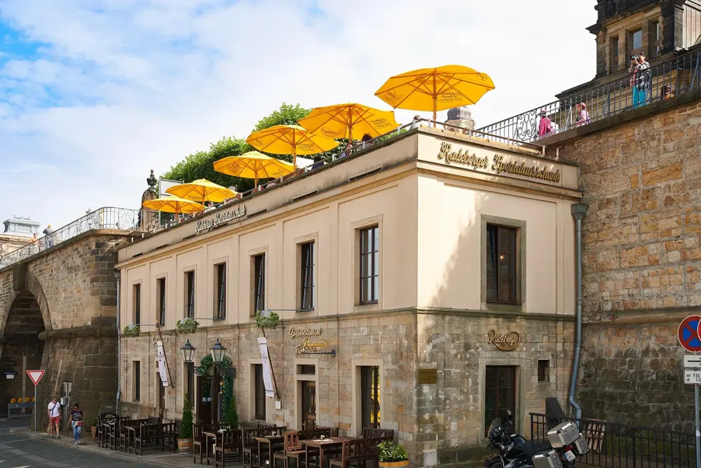 Building with terrace yellow umbrellas and Radeberger Spezialausschank sign. Stone wall and street with tables and people.