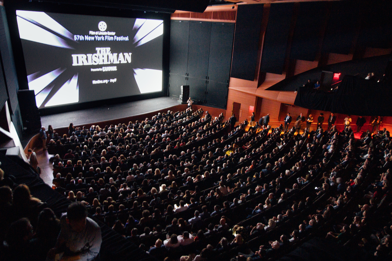 Una gran audiencia asiste al estreno de una película en un festival en un teatro moderno. El título de la película se muestra en una pantalla en el escenario.