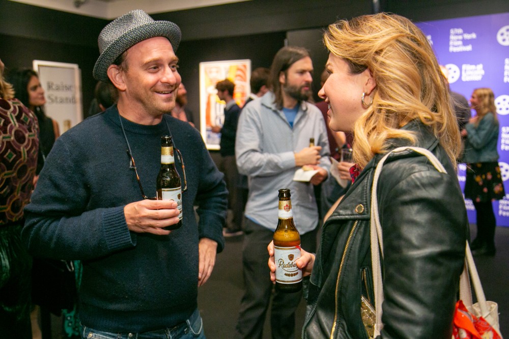 A man and woman talk and hold beer bottles at a crowded indoor event.