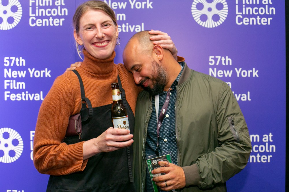 A man and a woman smile at an event; he holds a brochure, she holds a beer.